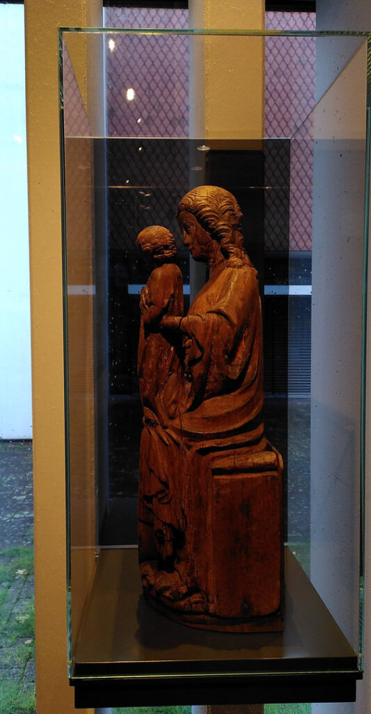 A photo of a wooden sculpture of a sitting woman holding up a child, in a glas case