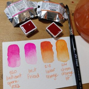A photo of watercolour half-pans, their packaging, a paper scrap with watercolour swatches, and a paintbrush