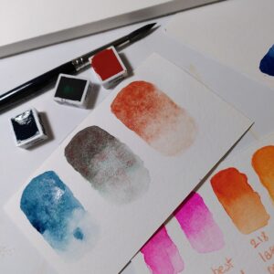 A photo of watercolour half-pans, paper scraps with watercolour swatches, and a paintbrush